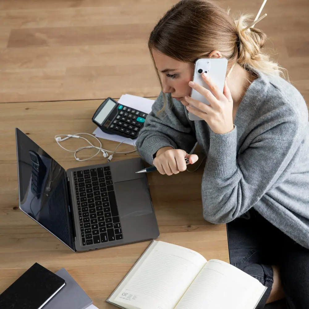 Woman reviewing finances with laptop, calculator, and phone — multitasking to manage monthly subscriptions.
