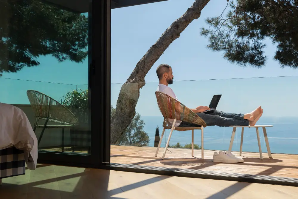 Man working remotely with a laptop on a balcony overlooking the ocean, representing digital nomad financial freedom.