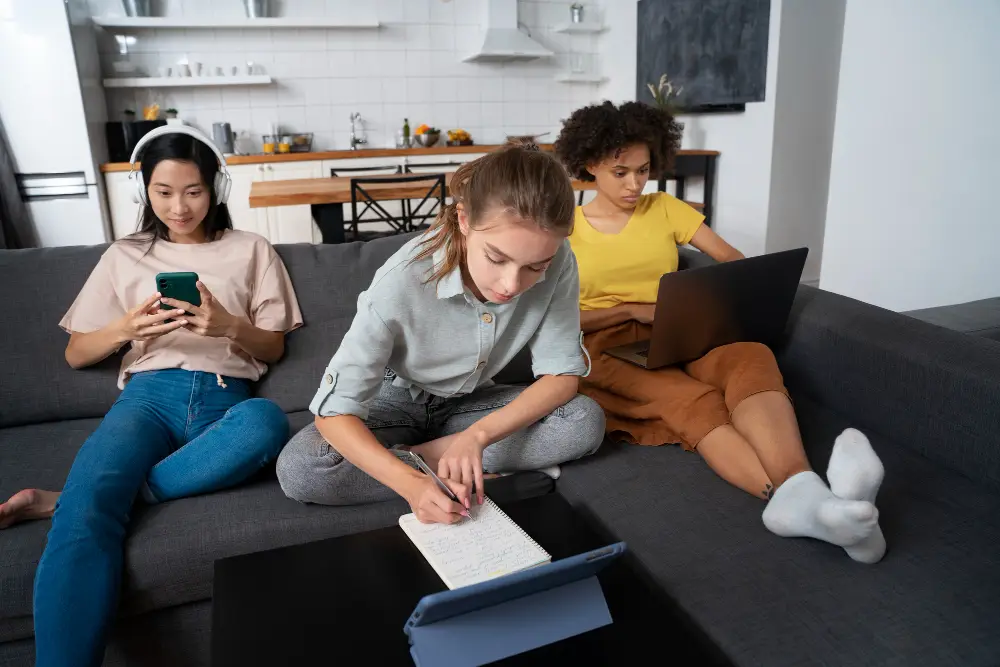Three young women sitting on a couch at home—one using a smartphone with headphones, one writing in a notebook, and one working on a laptop—representing Gen Z engaging in remote side hustles and digital learning.