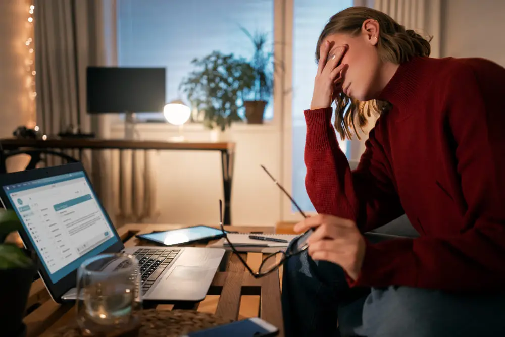 Woman overwhelmed and exhausted in front of a laptop at night, symbolizing side hustle fatigue and digital burnout in 2025.