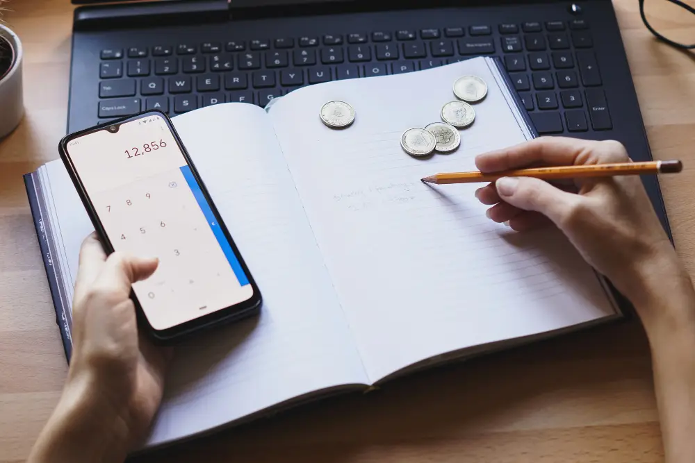 Person calculating monthly expenses with phone, notebook, and coins on a desk — showing personal finance planning.