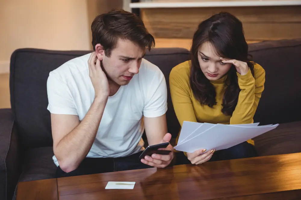 A young couple sits on a couch looking overwhelmed as they review bills and documents—symbolizing financial stress and money management challenges faced by Gen Z.