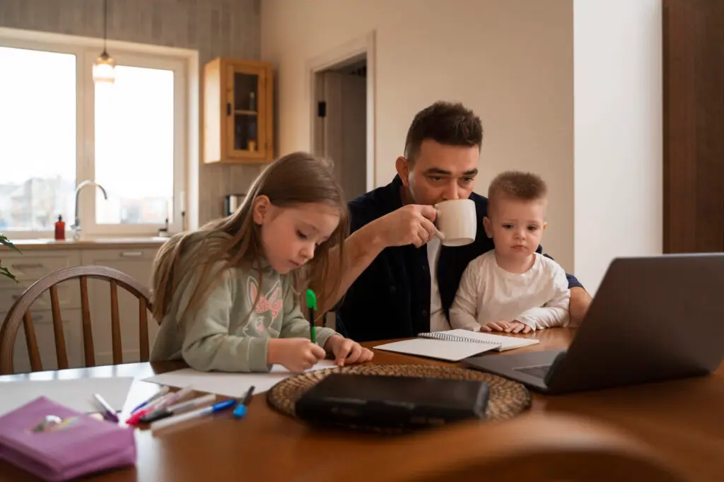 Father drinking coffee while working on a laptop with his two young children at the table, one drawing and the other watching.